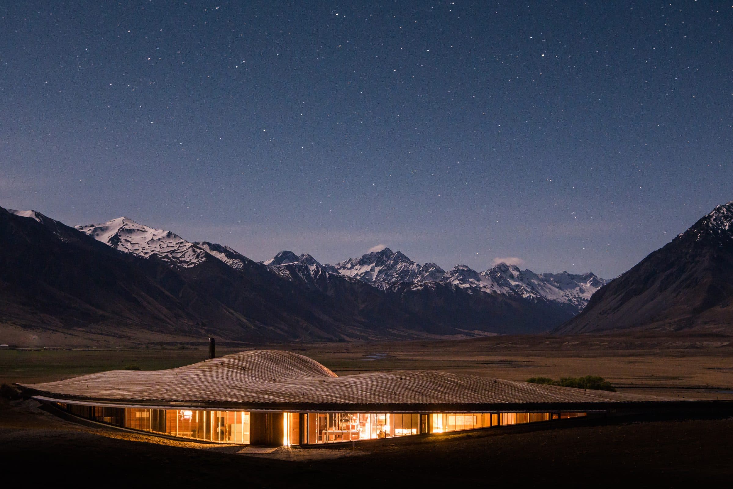 The Lindis lodge with its sculptural curved roof lit at dusk beneath snowed mountains
