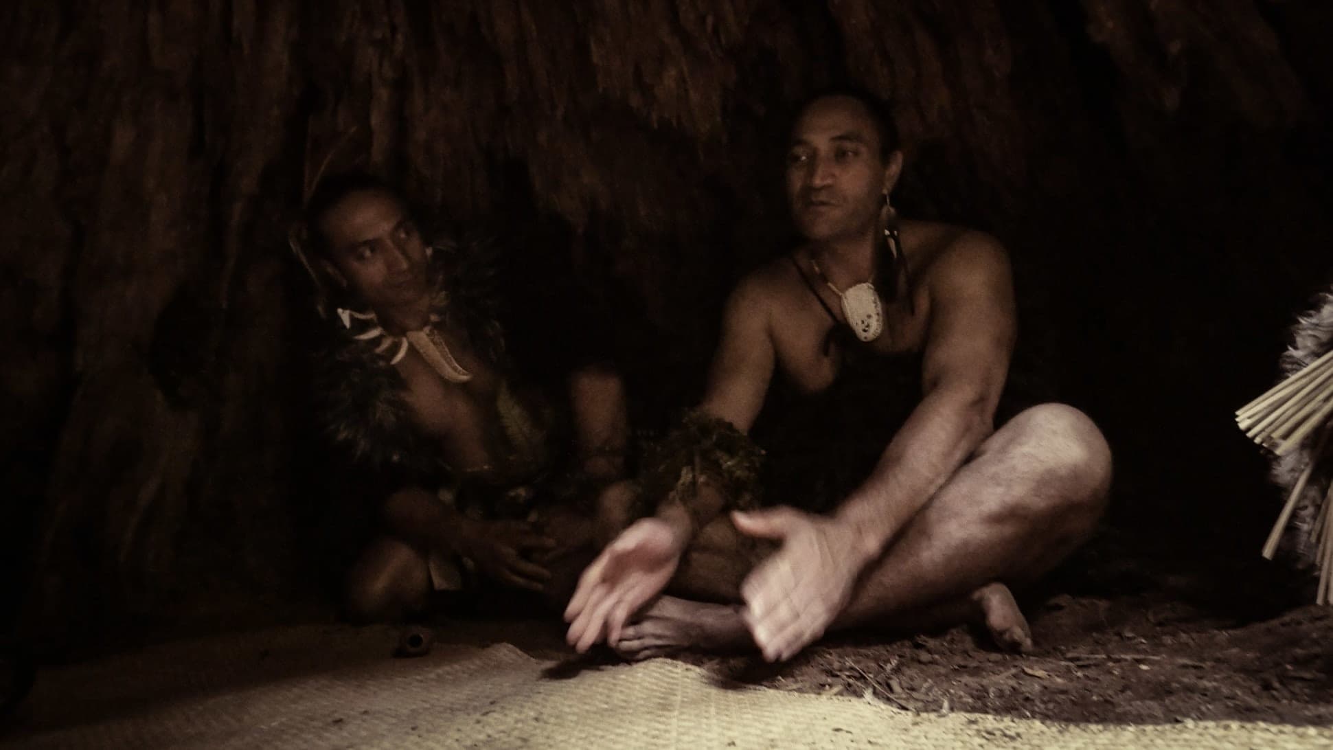 Two Māori hosts seated on a woven mat in dim light, one speaking